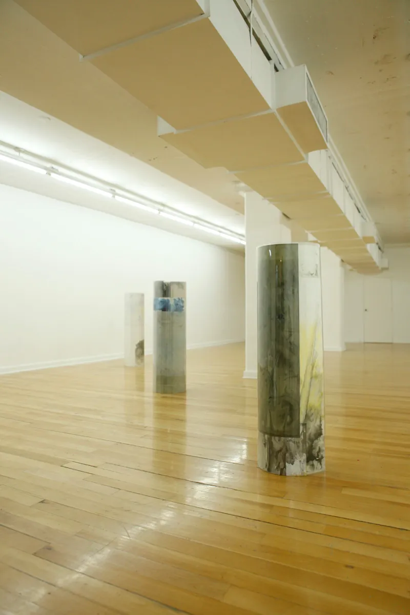 Sculpture installation where a series of drawings are rolled up between two polycarbonate sheets, creating a tube like shape standing vertically and aligned in a line, detail of three of them together, the show in general had a dimmed light set up