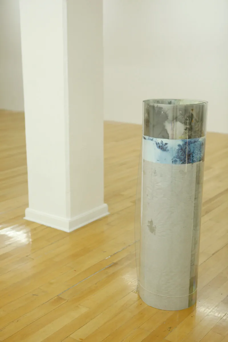 Sculpture installation where a series of drawings are rolled up between two polycarbonate sheets, creating a tube like shape standing vertically and aligned in a line, detail of only one of them, a blue grey marble like look can be visible partially from the drawings