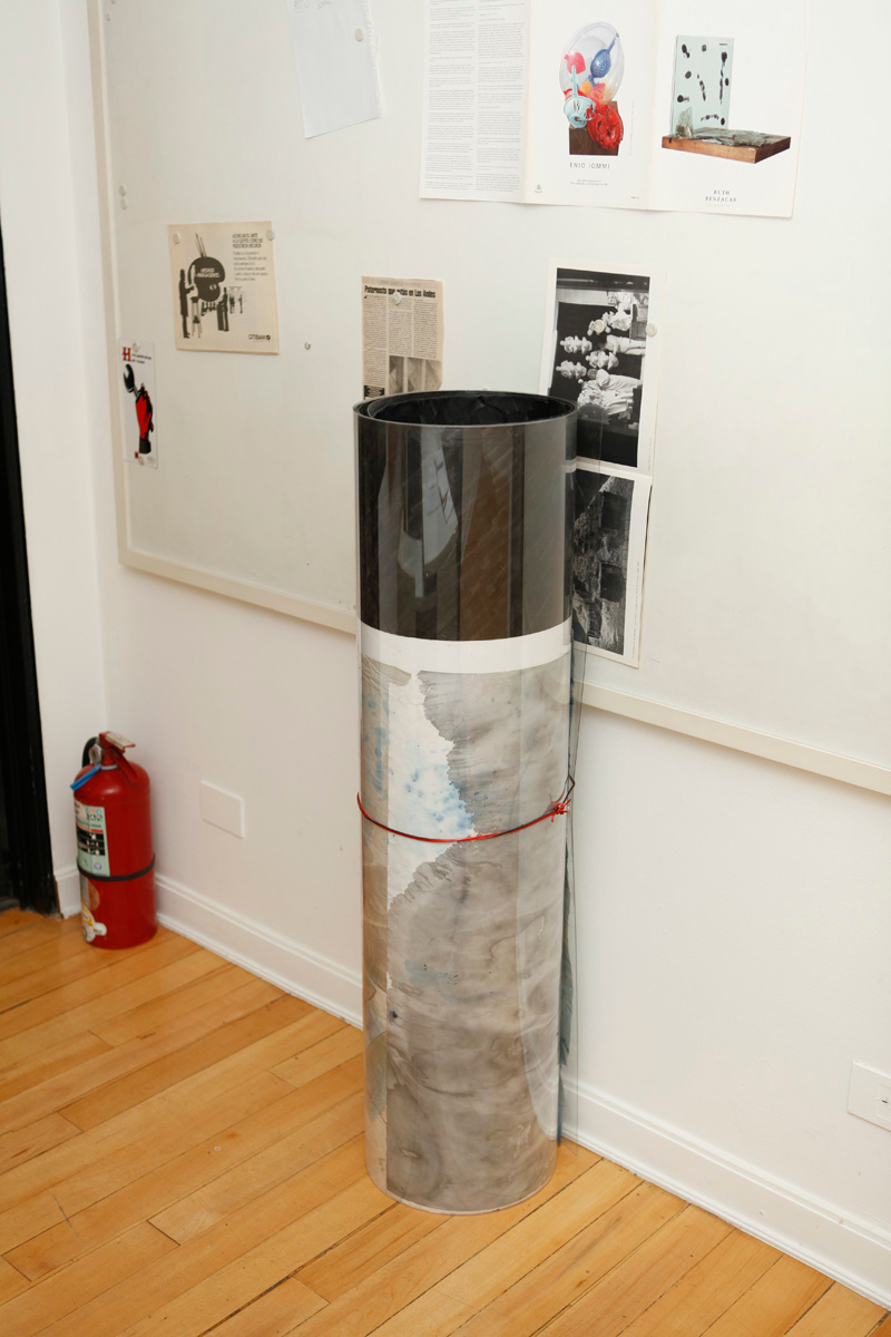 Sculpture installation where a series of drawings are rolled up between two polycarbonate sheets, creating a tube like shape standing vertically, detail of one of them, installed separately, where a set of posters from the gallery hanged from a metal board